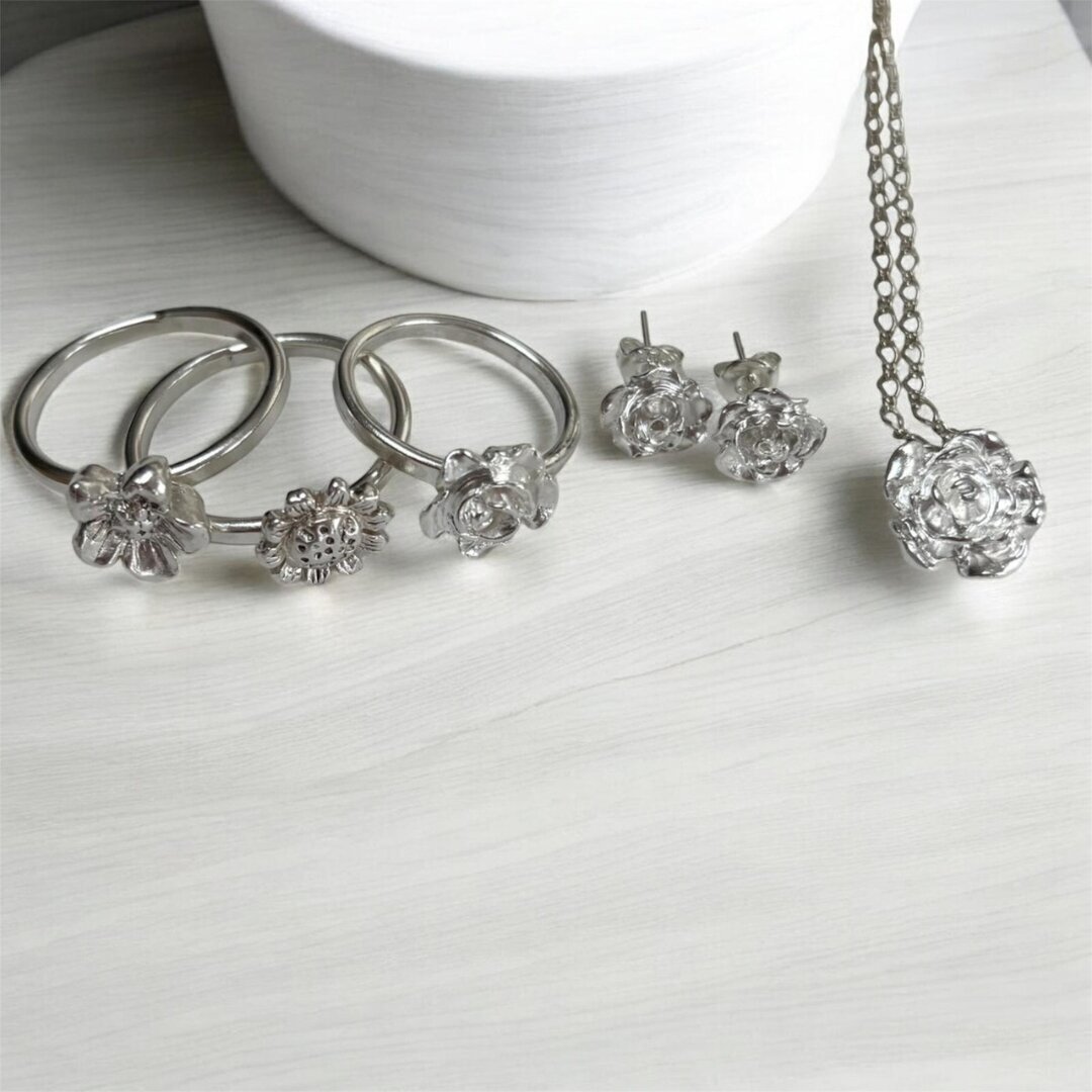Handcrafted silver jewellery including floral rings, stud earrings and a pendant necklace displayed on a white surface.